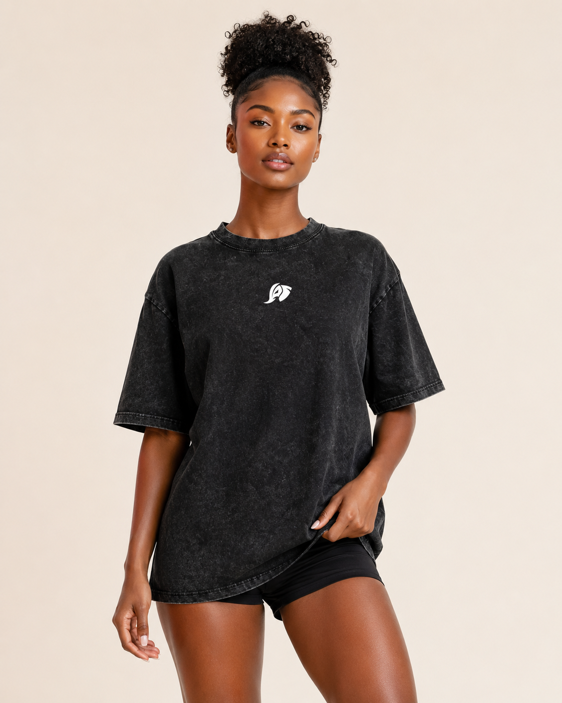 A woman modeling an oversized black acid-wash t-shirt with a small white graphic on the left chest, paired with black shorts, against a solid beige background.